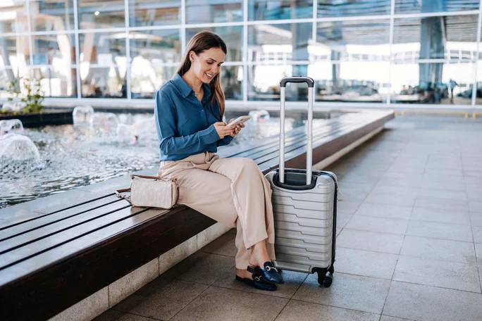 Imaginea prezintă o persoană așezată pe o bancă într-un spațiu public, posibil un aeroport. Persoana privește un telefon mobil și are o geantă mică alături pe bancă. Lângă aceasta se află un troler argintiu cu mânerul extins. În fundal se observă o fântână decorativă cu jeturi de apă și ferestre mari care reflectă interiorul clădirii.