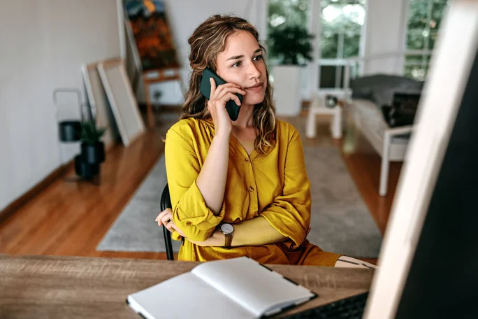 O femeie îmbrăcată într-o rochie galbenă este așezată la un birou, vorbind la telefon. Decorul pare a fi un birou de acasă modern, bine luminat, cu podea din lemn și un design minimalist. Pe birou se află un carnețel și un pix, iar în fundal se pot observa mobilier și opere de artă.