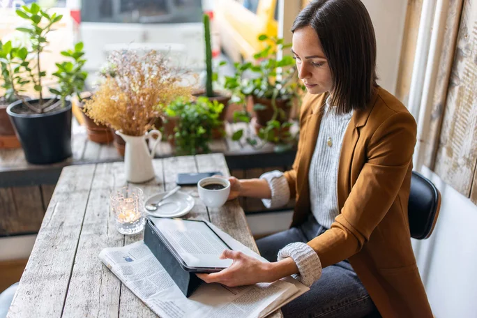 Imaginea surprinde o scenă relaxantă într-un spațiu confortabil, posibil o cafenea. Elementele vizibile includ:  O persoană așezată la o masă din lemn, ținând o ceașcă de cafea într-o mână Cu cealaltă mână, persoana citește de pe un dispozitiv electronic (probabil o tabletă sau un e-reader) aflat pe masă Pe masă și pe pervazul ferestrei se află plante în ghiveci și flori uscate, care contribuie la o atmosferă caldă și primitoare Sub dispozitivul electronic se află un ziar desfășurat parțial În fundal se vede o fereastră prin care se observă vehicule parcate afară Imaginea transmite o stare de liniște și concentrare, potrivită pentru lectură sau reflecție într-un mediu plăcut. 