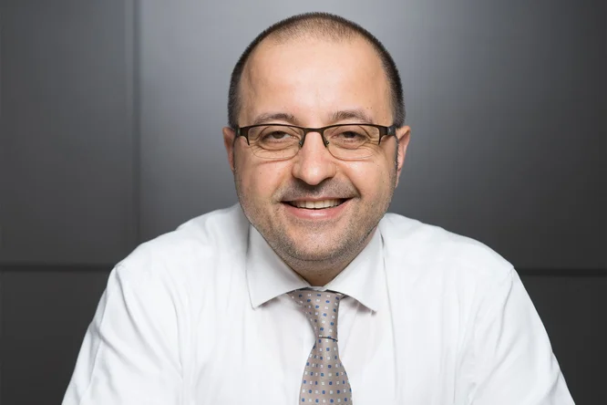 Un bărbat îmbrăcat într-o cămașă albă și o cravată cu model este așezat într-un spațiu interior, pe un fundal gri închis. Decorul pare profesional, cu un stil neutru și minimalist. Accentul este pus pe ținută și pe prezentarea formală în ansamblu.