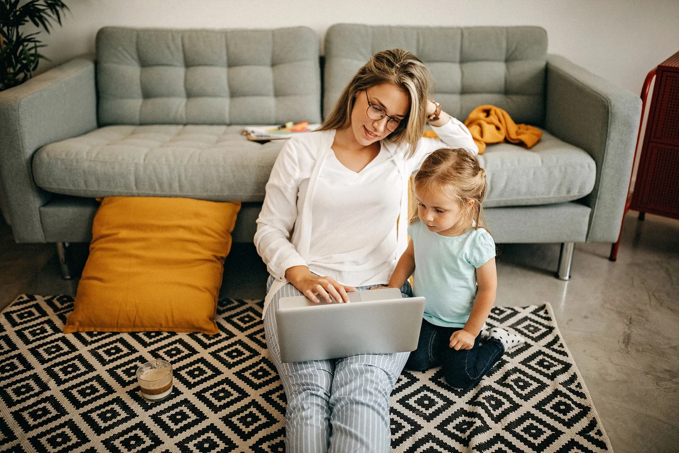 O mama si copilul ei mic stau pe un covor cu model in fata unei canapele gri. Mama foloseste un laptop in timp ce copilul priveste. In fundal sunt vizibile o perna galbena si o masa mica cu obiecte.