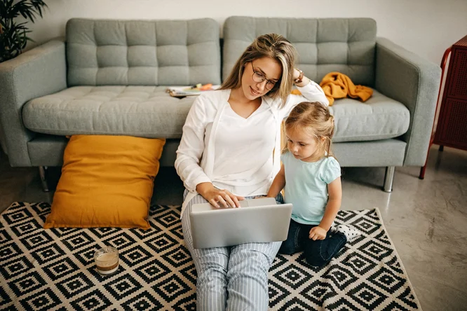 O mama si copilul ei mic stau pe un covor cu model in fata unei canapele gri. Mama foloseste un laptop in timp ce copilul priveste. In fundal sunt vizibile o perna galbena si o masa mica cu obiecte.