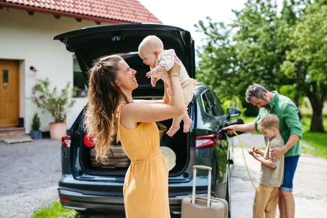 O familie se pregateste pentru o calatorie. O femeie in rochie galbena tine un bebelus langa portbagajul deschis al unei masini negre. Langa ea se afla o valiza cu maner extins. In fundal, un alt adult conecteaza un incarcator pentru vehicule electrice, in timp ce un copil se uita la ceva in mainile sale. Scena are loc afara, cu verdeata si o parte dintr-o casa vizibile.