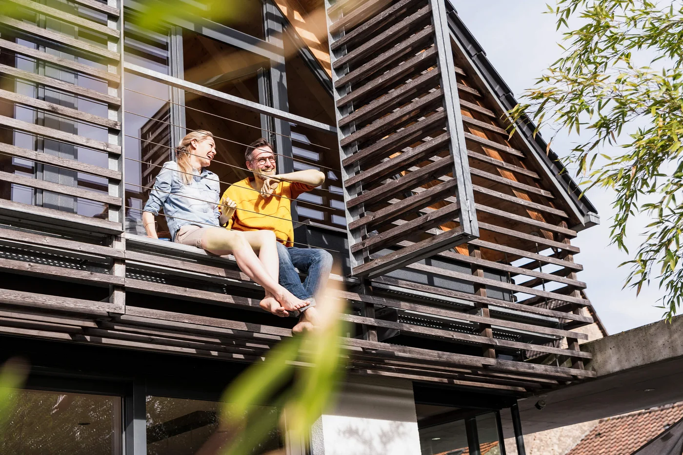 Doua persoane stau pe marginea unui balcon sau terase a unei cladiri moderne. Cladirea are ferestre mari si jaluzele orizontale. In prim-plan sunt vizibile cateva frunze verzi, sugerand ca sunt copaci sau plante in apropiere.