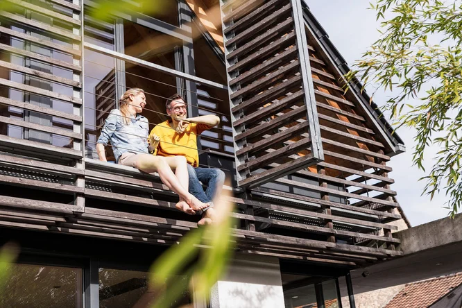 Doua persoane stau pe marginea unui balcon sau terase a unei cladiri moderne. Cladirea are ferestre mari si jaluzele orizontale. In prim-plan sunt vizibile cateva frunze verzi, sugerand ca sunt copaci sau plante in apropiere.