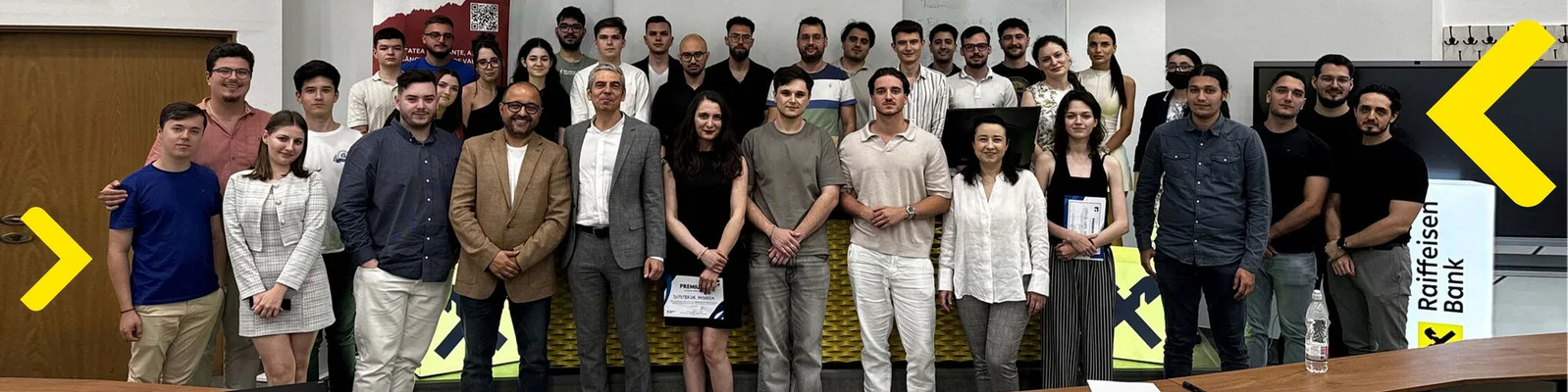 Un grup de aproximativ 25 de persoane sta impreuna intr-un cadru formal sau profesional. Persoanele sunt aranjate in trei randuri, iar unele dintre ele tin in mana certificate sau documente. In partea dreapta a imaginii este vizibil logo-ul Raiffeisen Bank, sugerand ca evenimentul este sponsorizat sau organizat de banca. In fundal se observa o usa din lemn in stanga si postere sau bannere in spatele grupului.