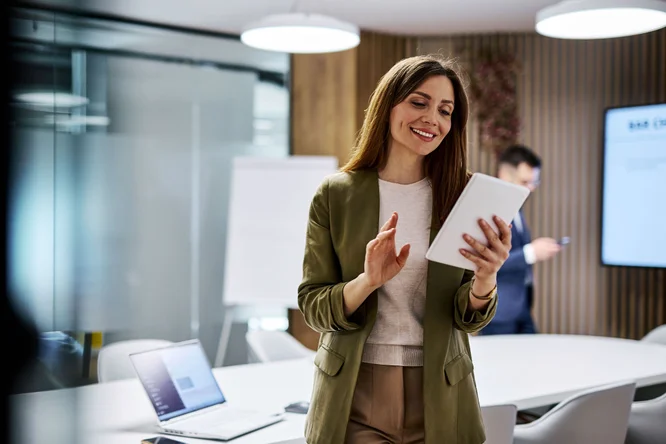 Imaginea prezintă o scenă într-un birou modern, cu următoarele elemente:  O persoană în ținută business casual (sacou verde peste o cămașă deschisă la culoare) care ține și interacționează cu o tabletă În fundal, o altă persoană folosește un smartphone Spațiul este amenajat cu mobilier contemporan: mese, scaune, panouri de sticlă, panouri de lemn pe pereți și corpuri de iluminat suspendate Pe o masă se află un laptop deschis În fundal se observă un whiteboard sau flipchart Pe perete este montat un ecran mare, care afișează conținut neclar în această imagine Atmosfera generală este una profesională, specifică unui mediu de lucru colaborativ sau unei sesiuni de prezentare/planificare. 