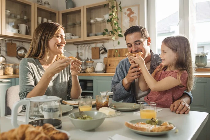 O familie se adună în jurul unei mese într-o bucătărie modernă și luminoasă pentru a savura micul dejun. Masa este aranjată cu croissante, suc de portocale, cafea și diverse creme tartinabile. Decorul include dulapuri din lemn deschis la culoare, ustensile suspendate și lumină naturală care pătrunde prin fereastră. Atmosfera este caldă și primitoare, cu un aer relaxat și degajat.