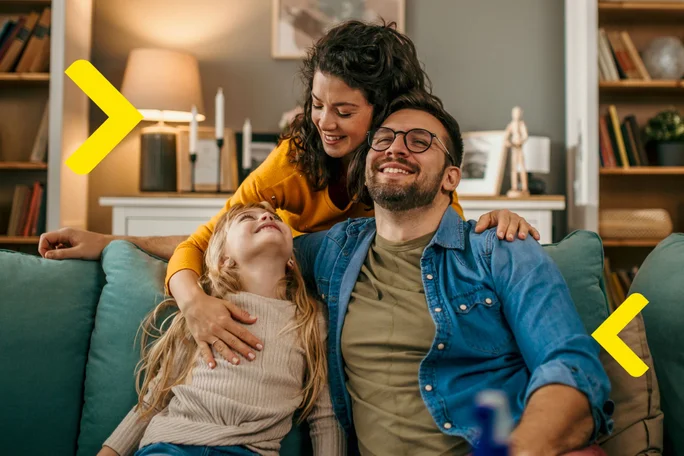 Trei persoane stau pe o canapea intr-un living. Persoana din stanga este un copil cu par blond lung, purtand un pulover bej si blugi albastri. Persoana din mijloc are fata ascunsa si imbratiseaza copilul cu un brat in jurul umerilor acestuia. Persoana din dreapta, de asemenea cu fata ascunsa, poarta o camasa de blugi peste un tricou verde masliniu si pantaloni rosii. In fundal, sunt rafturi pline cu carti si obiecte decorative, precum si o lampa pe o masa laterala care este aprinsa.