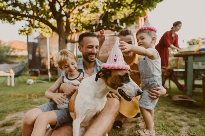 Familie cu copii si caine, posibil intr-o curte. Poarta palarii de petrecere si par sa sarbatoreasca un eveniment. In fundal se vad un copac si mobilier de exterior.