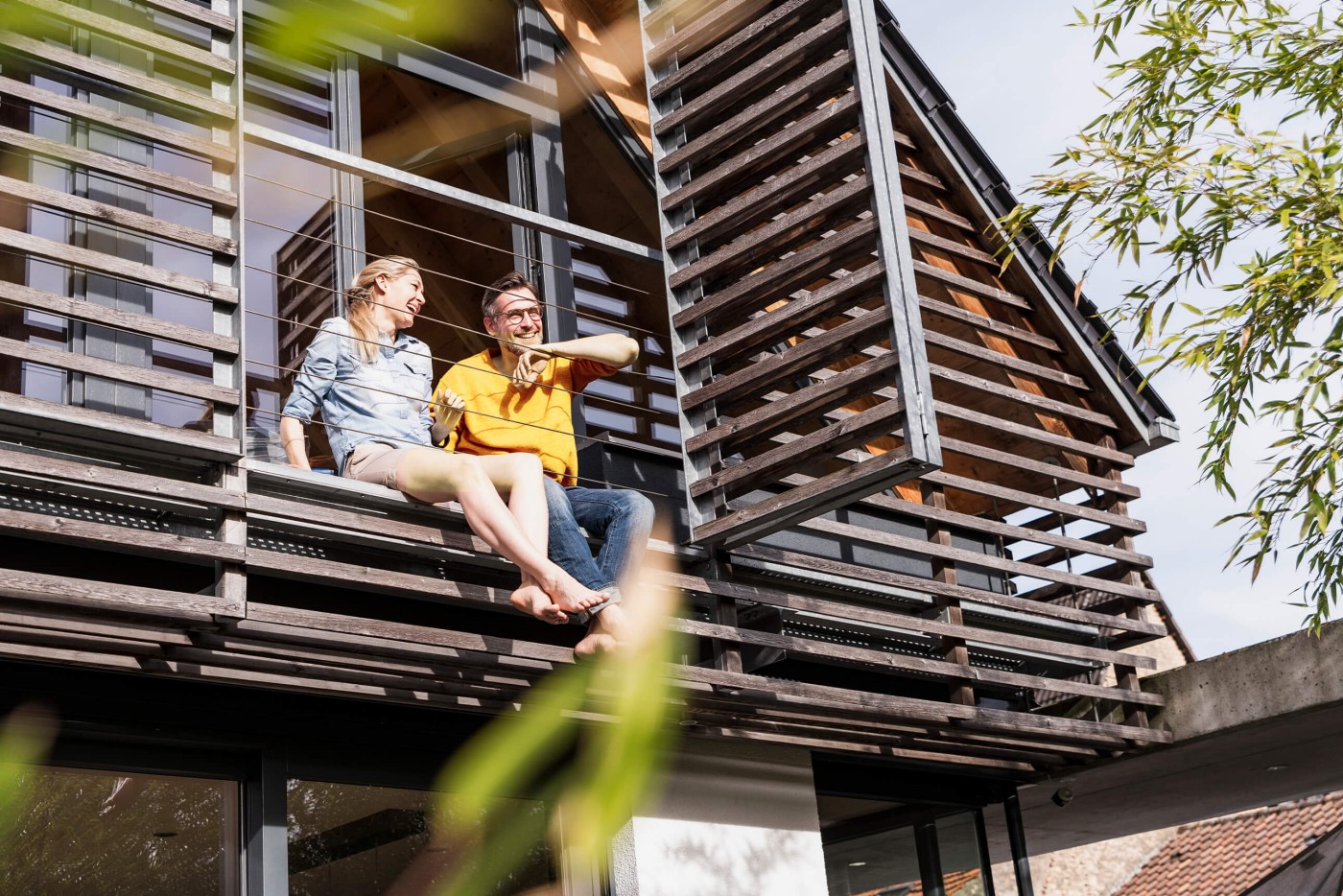 Doua persoane stau pe marginea unui balcon sau terase a unei cladiri moderne. Cladirea are ferestre mari si jaluzele orizontale. In prim-plan sunt vizibile cateva frunze verzi, sugerand ca sunt copaci sau plante in apropiere.