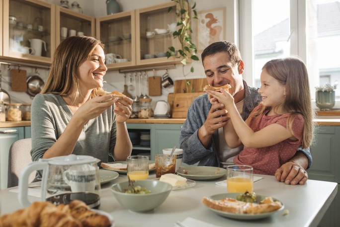 O familie se adună în jurul unei mese într-o bucătărie modernă și luminoasă pentru a savura micul dejun. Masa este aranjată cu croissante, suc de portocale, cafea și diverse creme tartinabile. Decorul include dulapuri din lemn deschis la culoare, ustensile suspendate și lumină naturală care pătrunde prin fereastră. Atmosfera este caldă și primitoare, cu un aer relaxat și degajat.