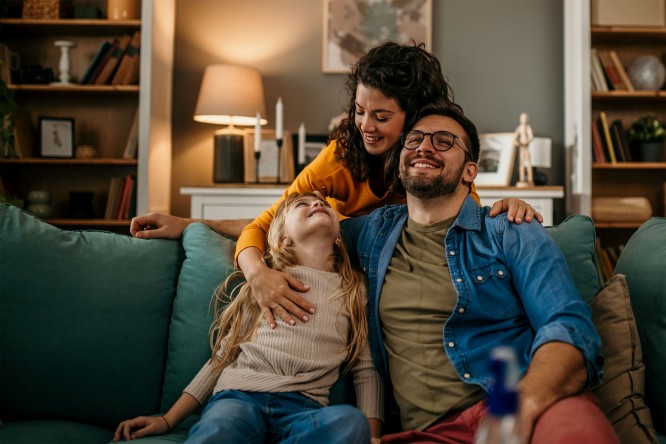 Trei persoane stau pe o canapea intr-un living. Persoana din stanga este un copil cu par blond lung, purtand un pulover bej si blugi albastri. Persoana din mijloc are fata ascunsa si imbratiseaza copilul cu un brat in jurul umerilor acestuia. Persoana din dreapta, de asemenea cu fata ascunsa, poarta o camasa de blugi peste un tricou verde masliniu si pantaloni rosii. In fundal, sunt rafturi pline cu carti si obiecte decorative, precum si o lampa pe o masa laterala care este aprinsa.