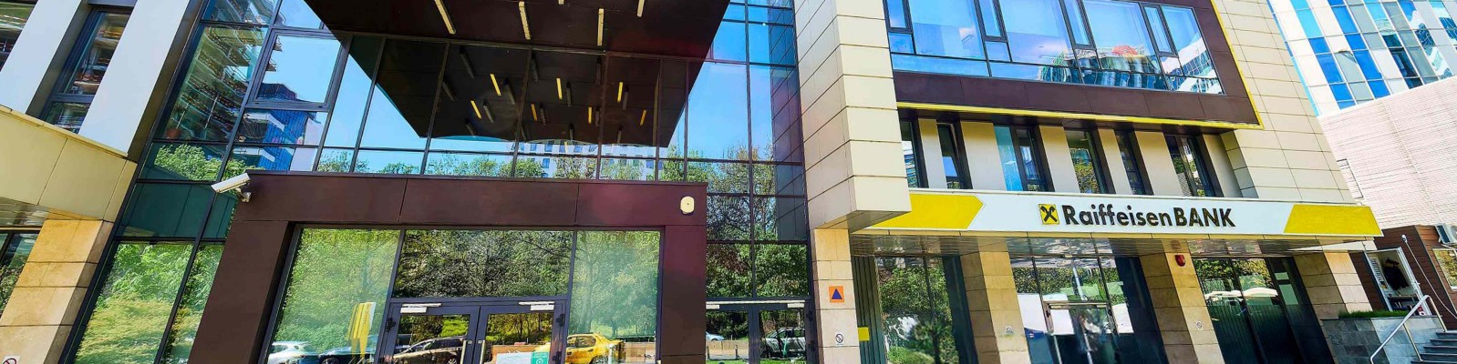 Imaginea prezintă exteriorul unei clădiri Raiffeisen Bank cu un design arhitectural contemporan. Decorul include ferestre din sticlă reflectorizantă și un mediu urban amenajat peisagistic. Numele băncii este vizibil clar pe panoul de semnalizare, însoțit de o paletă de culori galben și negru.